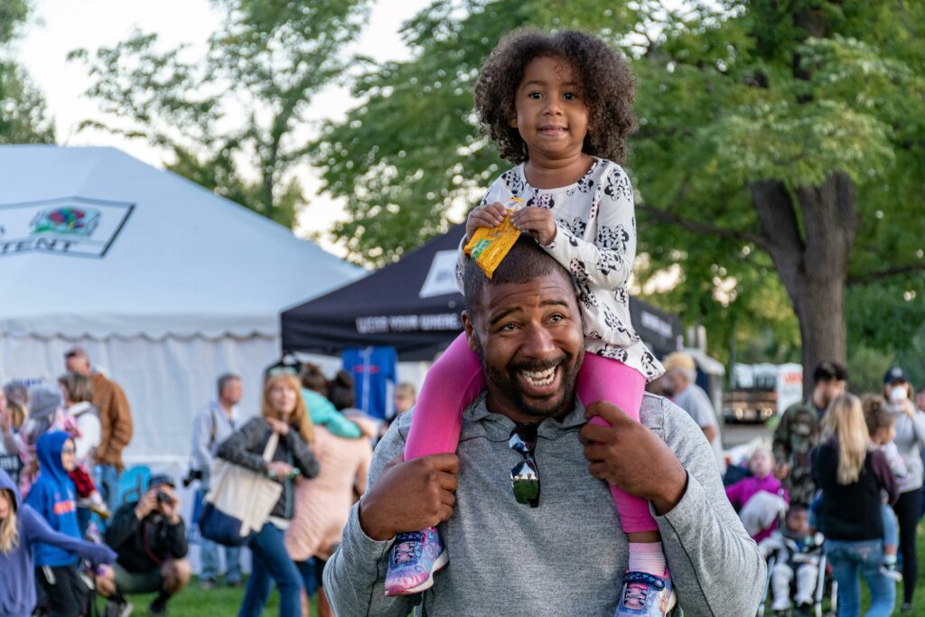 Young girl sitting on father's shoulders at outdoor family event