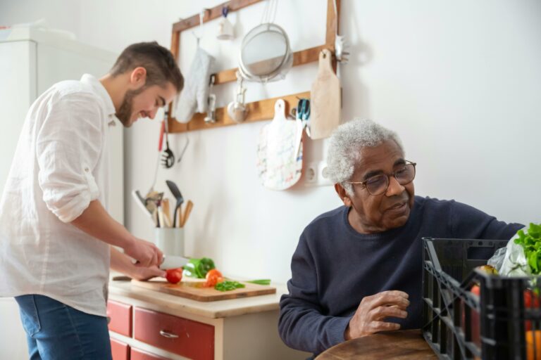 A young man in a kitchen prepares a meal for an elderly man sitting at a table, demonstrating volunteer efforts in Loudoun County’s Home Delivered Meals Program. This program connects volunteers with seniors who need assistance, offering critical support through meal preparation and delivery services.